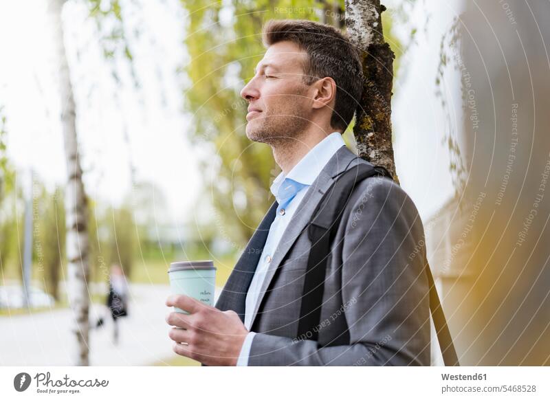 Entspannter Geschäftsmann mit Kaffee zum Mitnehmen in einem Park Birke Birken Betula Baum Bäume Baeume Laptoptasche Laptop-Tasche Reisende Reisender pendeln