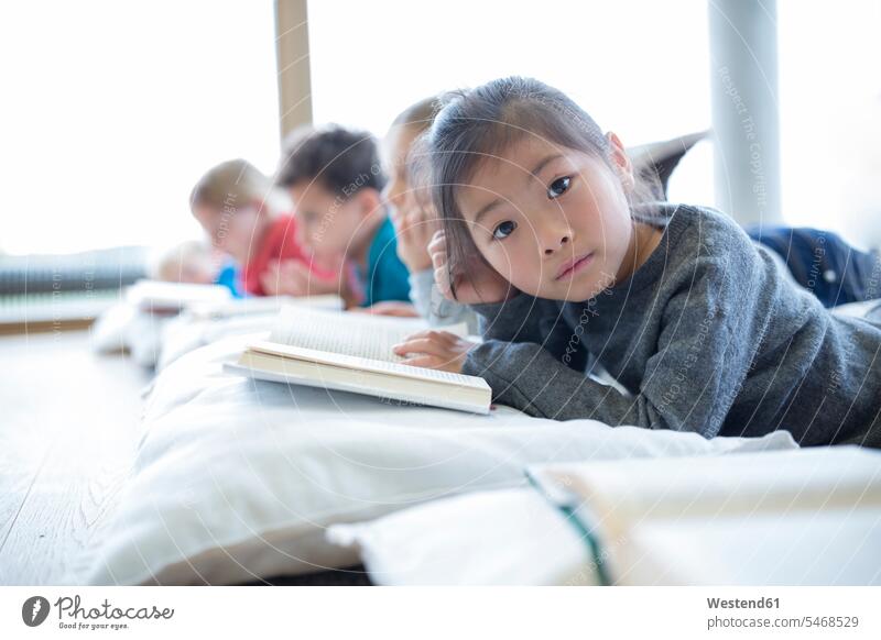 Porträt einer auf dem Boden liegenden Schülerin mit Klassenkameraden, die im Pausenraum der Schule ein Buch lesen Böden Boeden Lektüre Schulen Schuelerin