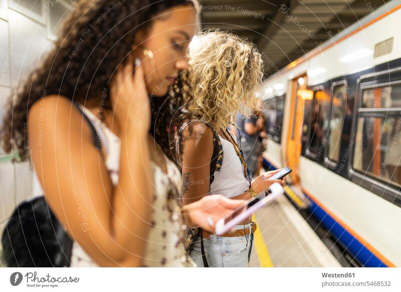 UK, London, zwei junge Frauen mit Handys warten am Bahnsteig einer U-Bahn-Station Freundinnen weiblich Mobiltelefon Handies Mobiltelefone Ubahn U-Bahnen