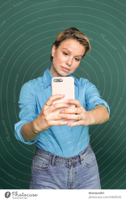 Porträt einer jungen Frau, die ein Selfie mit einem Smartphone vor einem grünen Hintergrund macht weiblich Frauen iPhone Smartphones Selfies Portrait Porträts
