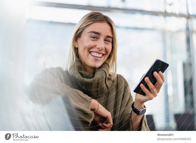 Porträt einer glücklichen jungen Frau, die mit einem Mobiltelefon im Wartebereich sitzt Telekommunikation telefonieren Handies Handys Mobiltelefone verreisen