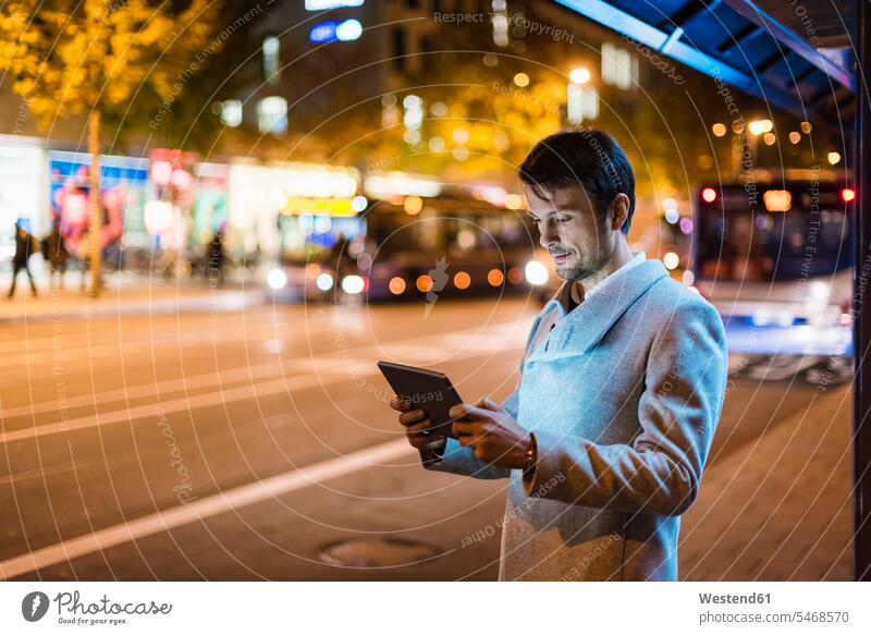 Geschäftsmann mit digitalem Tablet stehend an einer Bushaltestelle in der Nacht Stadt staedtisch städtisch Außenaufnahme draußen im Freien Europäer Kaukasier