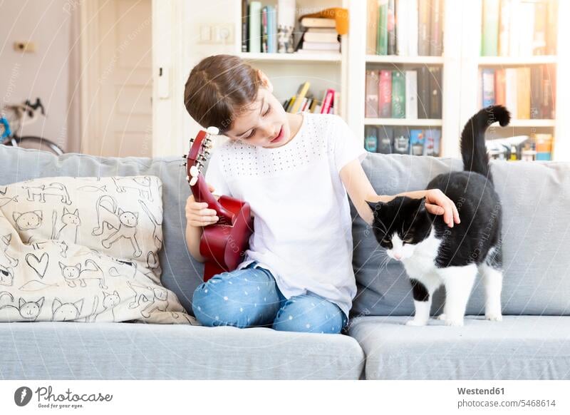 Mädchen mit Ukulele sitzt auf Couch und streichelt Katze Katzen sitzen sitzend Sofa Couches Liege Sofas weiblich Haustier Haustiere Tier Tierwelt Tiere Gitarre