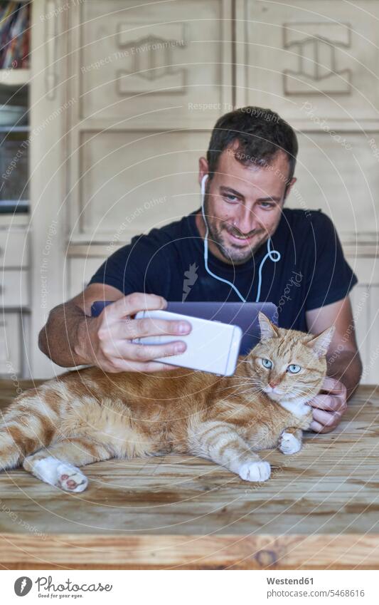 Mann sitzt am Tisch und fotografiert seine rothaarige Katze mit einem Smartphone Job Berufe Berufstätigkeit Beschäftigung Jobs Fotografen Photograph