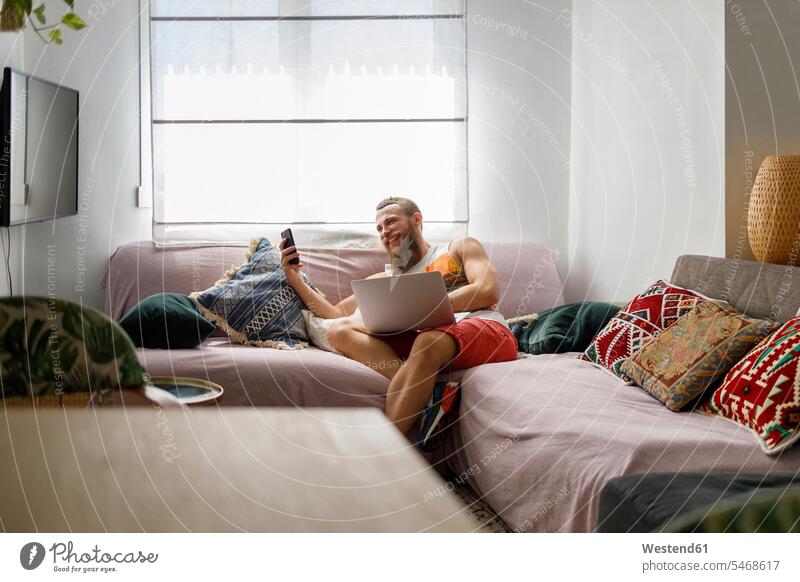 Lächelnder Mann benutzt Smartphone, während er mit Laptop auf dem Sofa im Wohnzimmer sitzt Farbaufnahme Farbe Farbfoto Farbphoto Innenaufnahme Innenaufnahmen