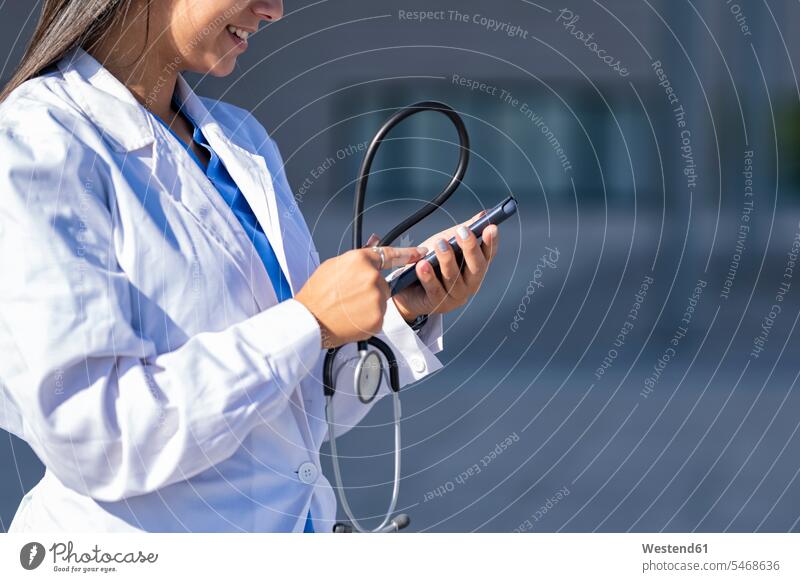 Arzt mit Stethoskop, der ein Mobiltelefon benutzt, während er gegen das Krankenhaus steht Farbaufnahme Farbe Farbfoto Farbphoto Außenaufnahme außen draußen