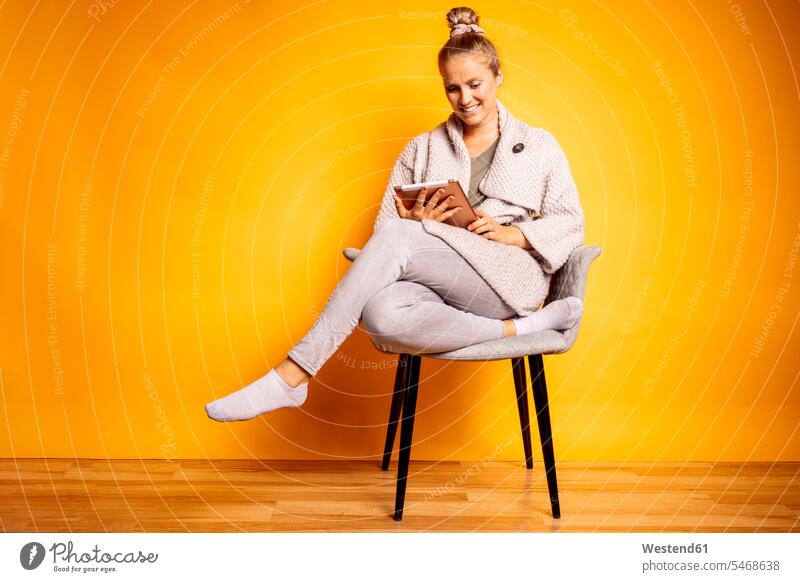 Lächelnde Frau benutzt digitales Tablett, während sie auf einem Stuhl vor gelbem Hintergrund sitzt Farbaufnahme Farbe Farbfoto Farbphoto farbiger Hintergrund