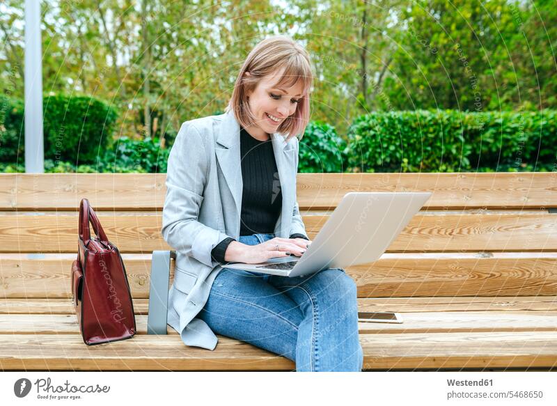 Lächelnde Frau sitzt auf Parkbank mit Laptop Job Berufe Berufstätigkeit Beschäftigung Jobs geschäftlich Geschäftsleben Geschäftswelt Geschäftsperson
