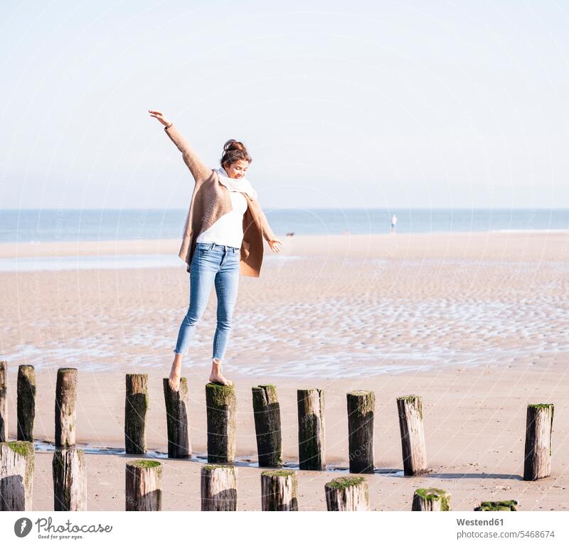 Lächelnde junge Frau mit ausgestreckten Armen, die an sonnigen Tagen am Strand auf Holzpfählen geht Farbaufnahme Farbe Farbfoto Farbphoto Niederlande Holland