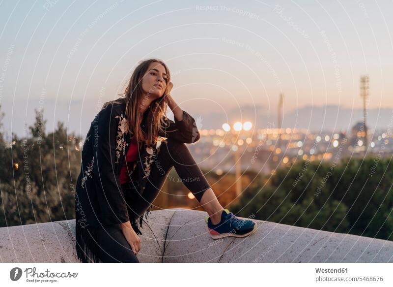 Spanien, Barcelona, Montjuic, junge Frau sitzt auf einer Mauer in der Abenddämmerung mit Stadt Lichter im Hintergrund Mauern weiblich Frauen stimmungsvoll