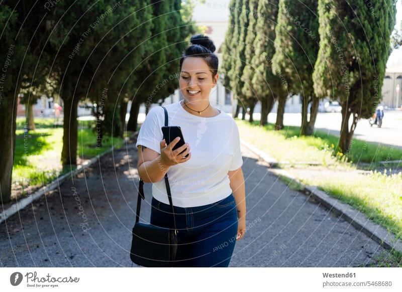 Lächelnde kurvenreiche junge Frau benutzt Mobiltelefon in einem öffentlichen Park Leute Menschen People Person Personen erwachsen Millennial 20 - 30 Jahre