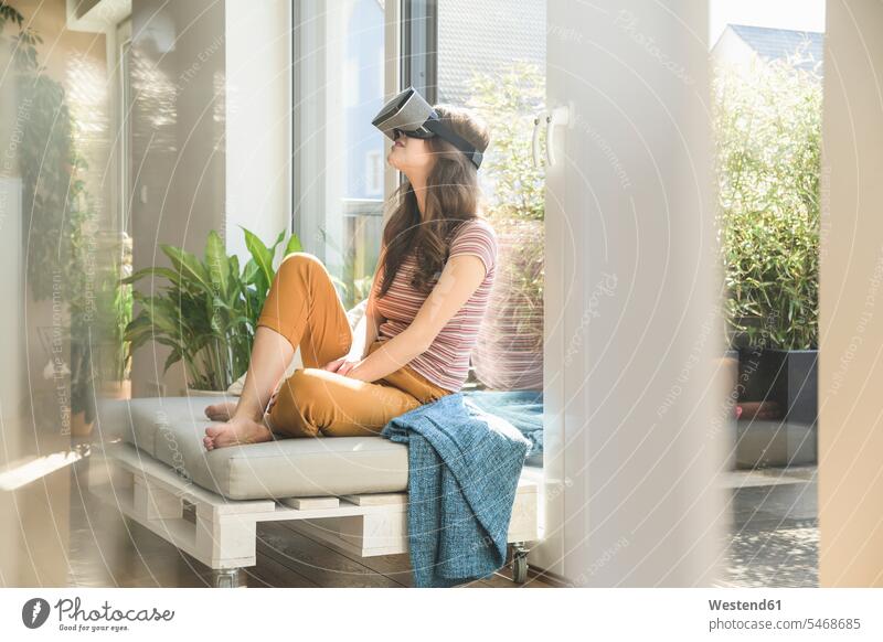 Junge Frau sitzt am Fenster zu Hause mit VR-Brille weiblich Frauen Zuhause daheim virtuell Virtualität sitzen sitzend Brillen Erwachsener erwachsen Mensch