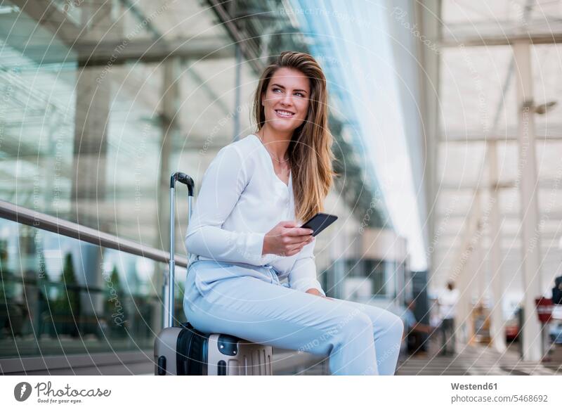 Lächelnde junge Geschäftsfrau sitzt auf Gepäck mit Handy und schaut sich um sitzen sitzend umherblicken umschauen umsehen Reisegepäck Mobiltelefon Handies