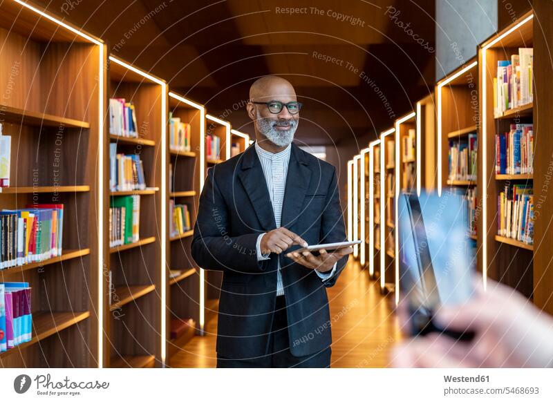 Porträt eines lächelnden Geschäftsmannes mit digitalem Tablet in einer Bibliothek Businessmann Businessmänner Geschäftsmänner Portrait Porträts Portraits Mann