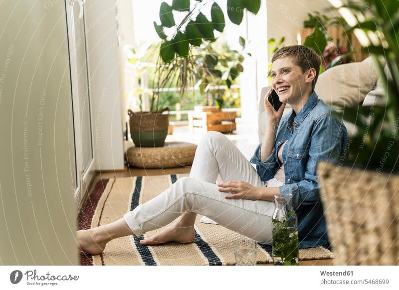 Fröhliche Frau spricht über Smartphone, während sie zu Hause auf dem Teppich sitzt Farbaufnahme Farbe Farbfoto Farbphoto Deutschland Freizeitbeschäftigung Muße