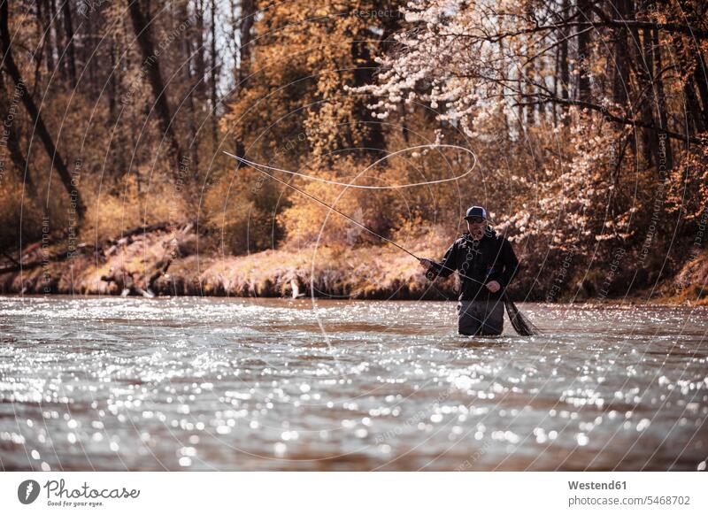 Fliegenfischer wirft Angelschnur aus, während er im Fluss am Wald steht Farbaufnahme Farbe Farbfoto Farbphoto Außenaufnahme außen draußen im Freien Fischer