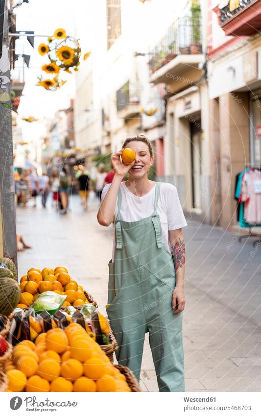 Porträt einer lachenden jungen Frau auf der Einkaufsstraße, die ihr Auge mit einer Orange bedeckt Leute Menschen People Person Personen Europäisch Kaukasier