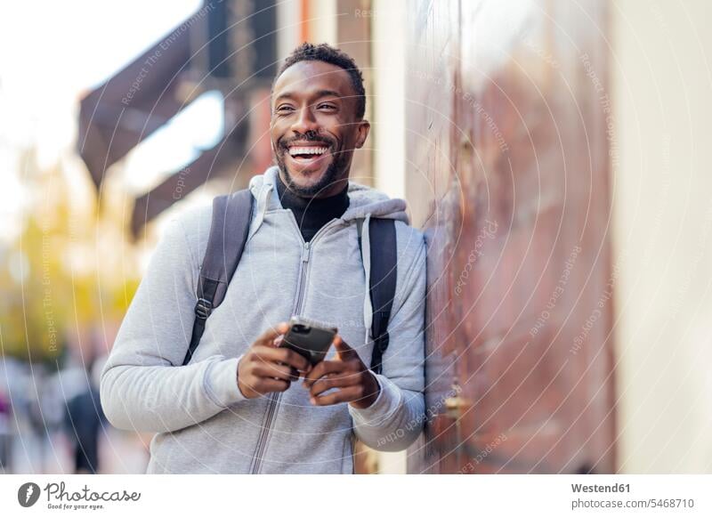 Lächelnder Mann, der ein Mobiltelefon benutzt, während er sich an die Wand lehnt Farbaufnahme Farbe Farbfoto Farbphoto Außenaufnahme außen draußen im Freien Tag