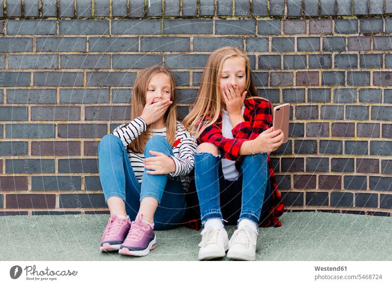 Porträt von zwei Mädchen, die vor einer Backsteinmauer sitzen und ein Selfie mit ihrem Smartphone machen iPhone Smartphones Selfies weiblich Backsteinwand