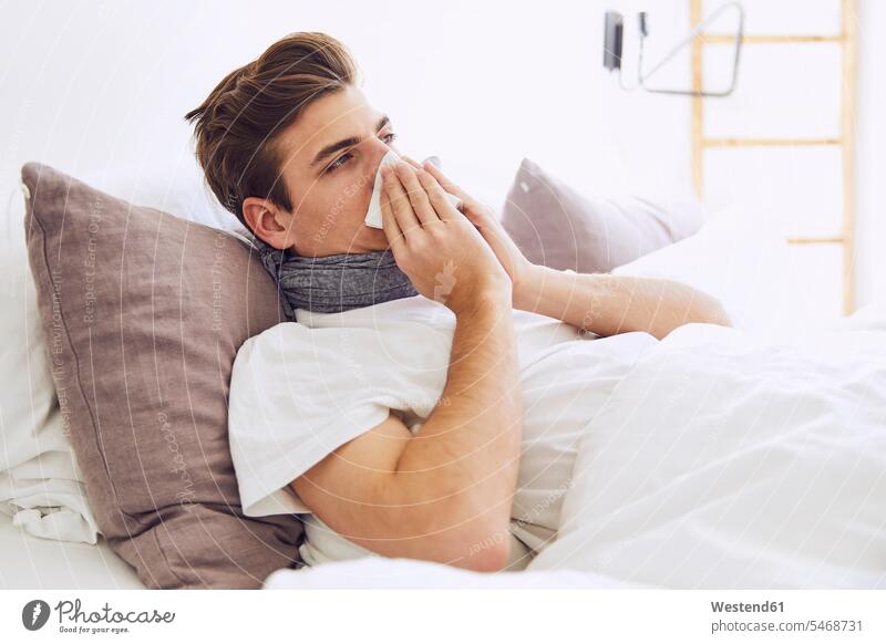 Kranker junger Mann schnäuzt sich die Nase, während er zu Hause auf dem Bett liegt Farbaufnahme Farbe Farbfoto Farbphoto Deutschland Innenaufnahme
