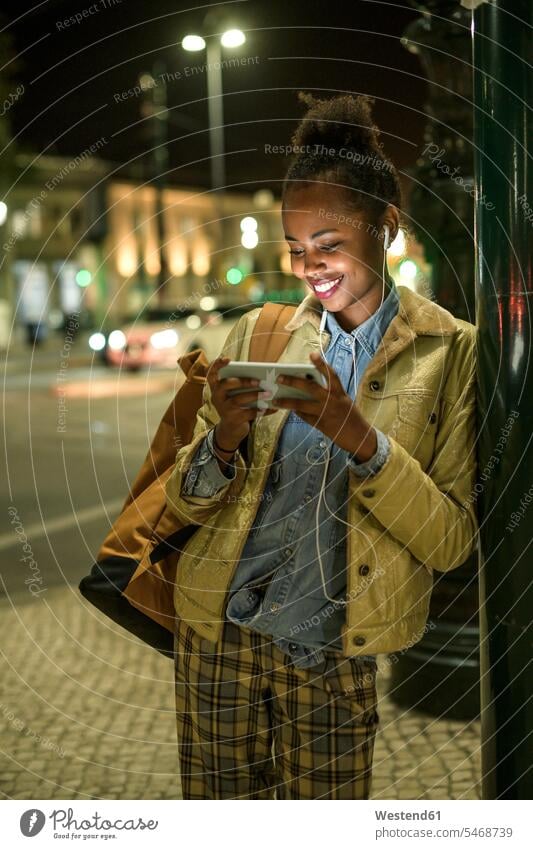 Porträt einer lächelnden jungen Frau mit Kopfhörer und Smartphone in der Stadt bei Nacht, Lissabon, Portugal Rucksäcke KFZ Verkehrsmittel Automobil Autos PKW