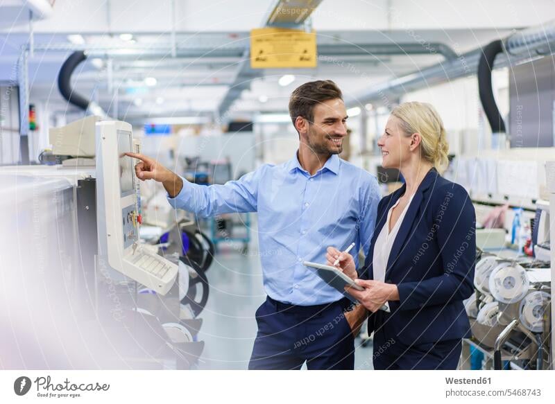 Lächelnder männlicher Techniker schaut eine Geschäftsfrau an, während er an einer Maschine in einer beleuchteten Fabrik steht Farbaufnahme Farbe Farbfoto