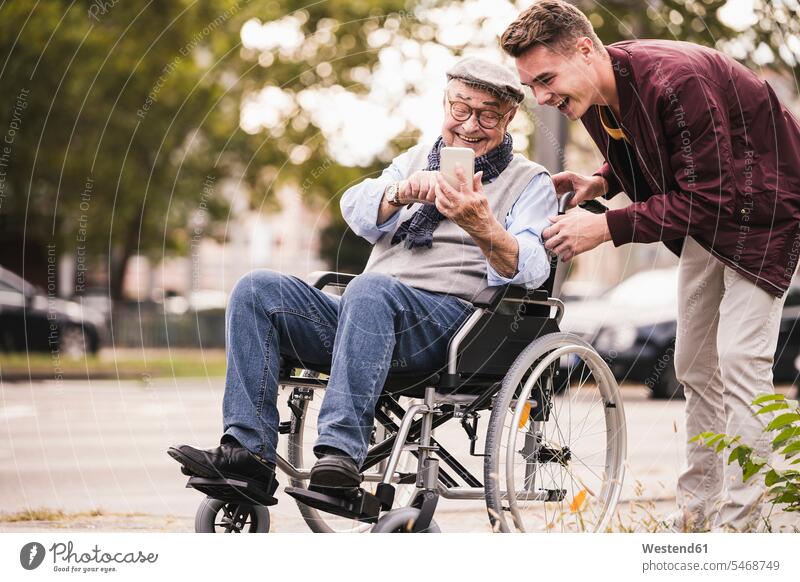 Lachender älterer Mann im Rollstuhl und sein erwachsener Enkel schauen gemeinsam auf Smartphone und haben Spaß Leute Menschen People Person Personen Europäisch