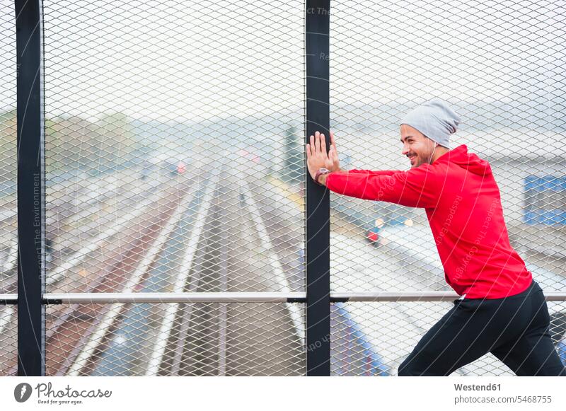 Junger Mann streckt sich auf einer Eisenbahnbrücke Gitterrost Raster Rost hoeren ausüben trainieren Übung freuen Farben Farbtoene Farbton Farbtöne rote roter