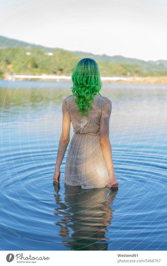 Rückenansicht einer jungen Frau mit gefärbten grünen Haaren, die im Wasser eines Sees steht stehen stehend weiblich Frauen angezogen faerben färben gefaerbt