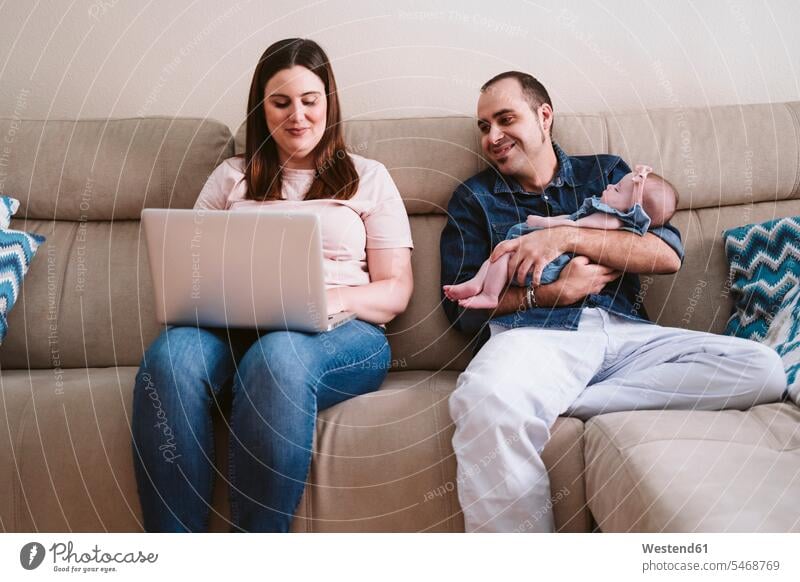 Vater trägt schlafende Tochter, während er die Frau am Laptop auf dem Sofa anschaut Farbaufnahme Farbe Farbfoto Farbphoto Innenaufnahme Innenaufnahmen innen