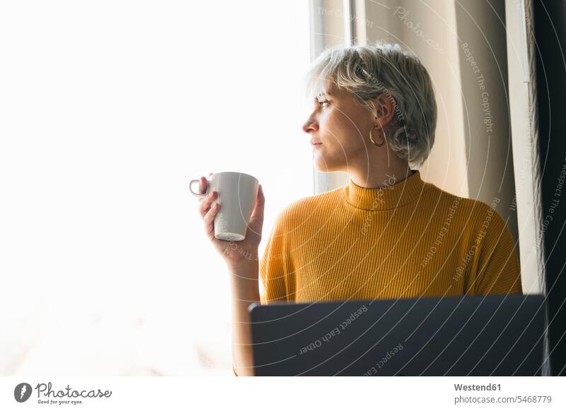 Frau schaut aus dem Fenster zu Hause mit Laptop und Kaffeetasse Leute Menschen People Person Personen Alleinstehende Alleinstehender Singles Unverheiratete