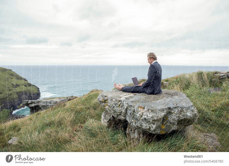 UK, Cornwall, Tintagel, Geschäftsmann sitzt mit Laptop auf einem Felsen an der Küste Businessmann Businessmänner Geschäftsmänner Notebook Laptops Notebooks