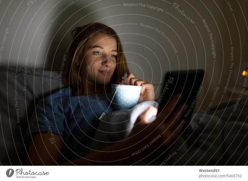 Junge Frau liegt zu Hause im Bett und trinkt abends eine Tasse Tee mit Tablette Leute Menschen People Person Personen Europäisch Kaukasier kaukasisch 1 Ein
