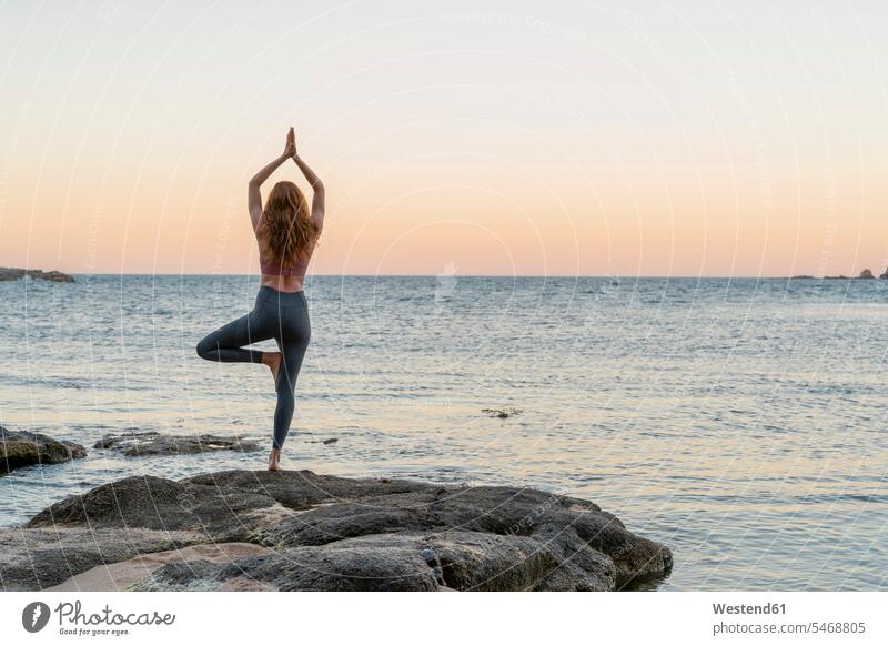 Junge Frau praktiziert Yoga am Strand, in Baumpose, während des Sonnenuntergangs am ruhigen Strand, Costa Brava, Spanien Leute Menschen People Person Personen
