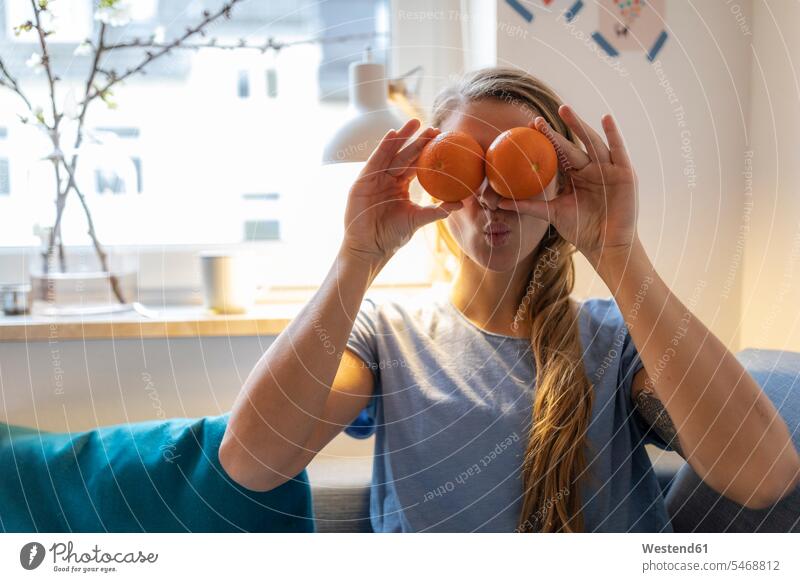 Verspielte junge Frau, die zu Hause ihre Augen mit Orangen bedeckt Leute Menschen People Person Personen Alleinstehende Alleinstehender Singles Unverheiratete