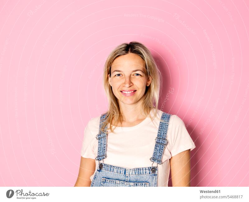Porträt einer lächelnden blonden Frau in Latzhose vor einem rosa Hintergrund blonde Haare blondes Haar Portrait Porträts Portraits weiblich Frauen Mensch