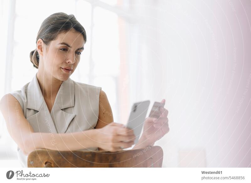 Geschäftsfrau, die eine Kreditkarte besitzt, während sie im Büro ein Mobiltelefon benutzt Farbaufnahme Farbe Farbfoto Farbphoto Innenaufnahme Innenaufnahmen