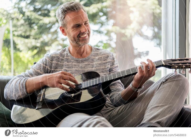 Glücklicher reifer Mann liegt zu Hause auf dem Sofa und spielt Gitarre Zuhause daheim spielen Gitarren lächeln sitzen sitzend sitzt Couches Liege Sofas Männer