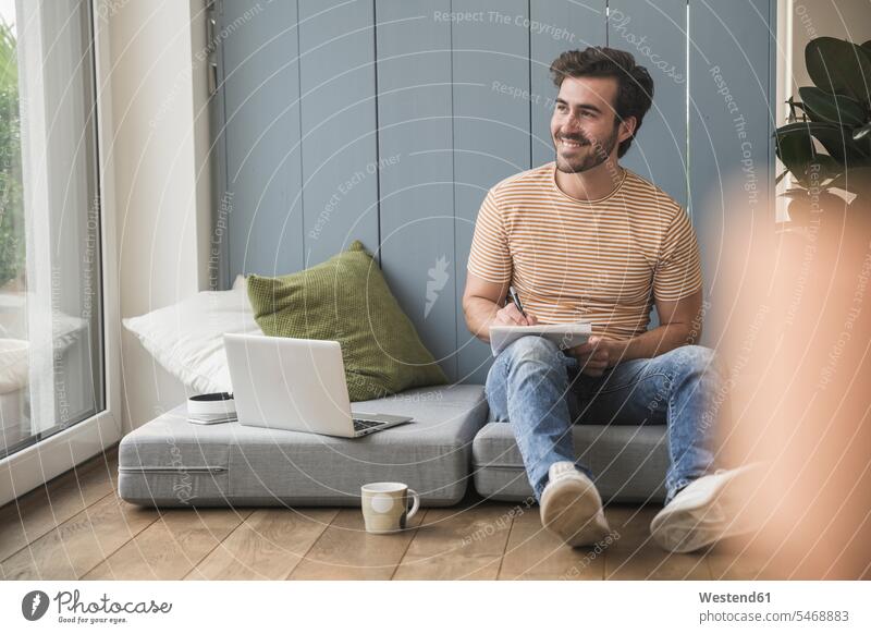 Junger Mann sitzt auf einer Matratze, benutzt einen Laptop und macht sich Notizen Deutschland Notizblock Notizbloecke Notizblöcke offenes Lächeln lachen