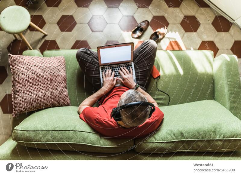 Reifer Mann sitzt auf dem Sofa und benutzt den Laptop, hört Musik Job Berufe Berufstätigkeit Beschäftigung Jobs Polster Couch Couches Liege Sofas Kopfhoerer