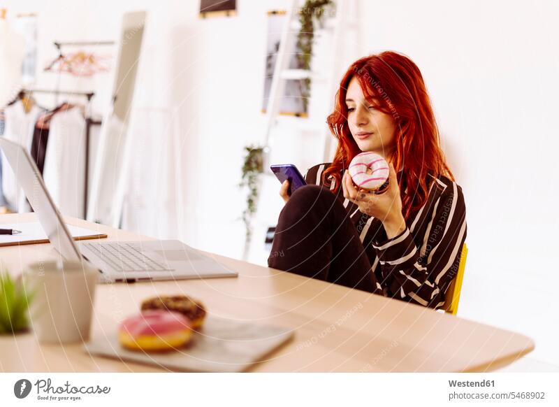Junge Geschäftsfrau hält Doughnut in der Hand, während sie im Büro sitzt und ihr Mobiltelefon benutzt Farbaufnahme Farbe Farbfoto Farbphoto Innenaufnahme