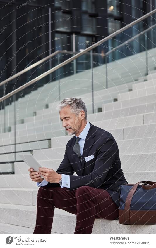 Modischer Geschäftsmann mit Reisetasche auf der Treppe sitzend mit Tablet Businessmann Businessmänner Geschäftsmänner modisch Treppenaufgang Tablet Computer