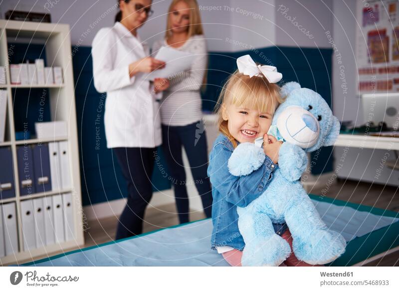 Mädchen umarmt Teddy in medizinischer Praxis mit Arzt und Mutter im Hintergrund Teddies weiblich Ärztin Aerztin Ärztinnen Doktorinnen Aerztinnen Mami Mutti