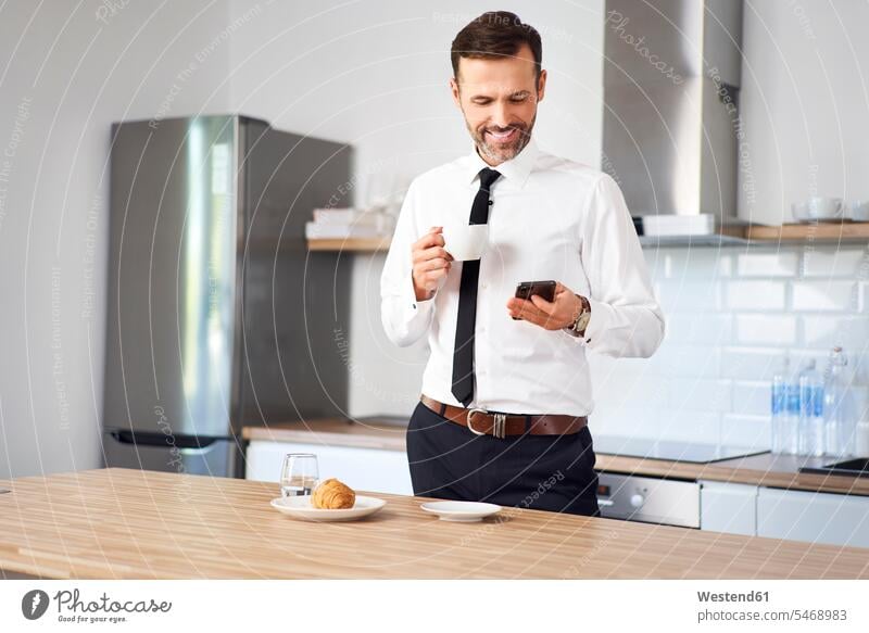 Mann steht in der Küche in der mit Kaffee und Blick auf sein Telefon vor der Arbeit trinken benutzen benützen Smartphone iPhone Smartphones morgens Morgen früh