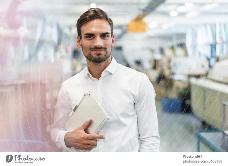 Selbstbewusster junger Ingenieur hält digitales Tablett in der Hand, während er in der beleuchteten Fabrik steht Farbaufnahme Farbe Farbfoto Farbphoto