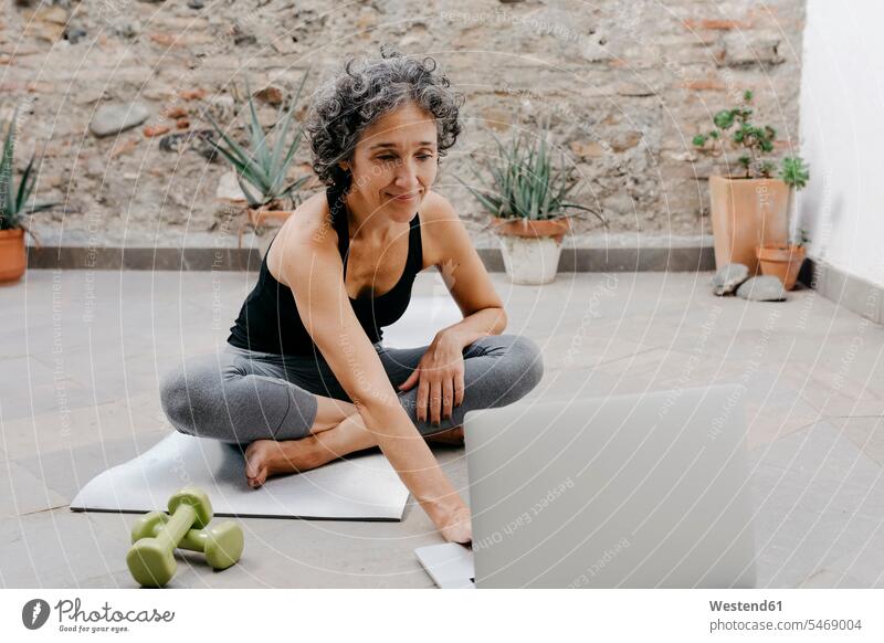 Lächelnde Frau benutzt Laptop zum Yoga-Lernen durch Online-Tutorial, während sie im Hinterhof sitzt Farbaufnahme Farbe Farbfoto Farbphoto Spanien Außenaufnahme