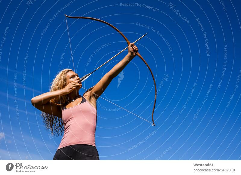 Bogenschützin, die mit dem Bogen gegen den blauen Himmel zielt zielen zielend Bogenschützinnen Bogenschuetzin Bogenschuetzinnen Boegen Bögen Bogenschütze