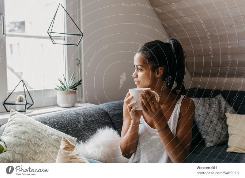 Junge Frau mit Tasse Kaffee sitzt auf der Couch zu Hause Blick aus dem Fenster Kaffeetasse Kaffeetassen weiblich Frauen Zuhause daheim sitzen sitzend Sofa