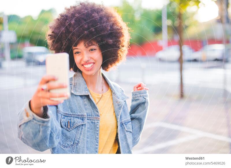 Glückliche junge Frau mit Afrofrisur macht ein Selfie in der Stadt Leute Menschen People Person Personen gelockt gelockte Haare gelocktes Haar lockig