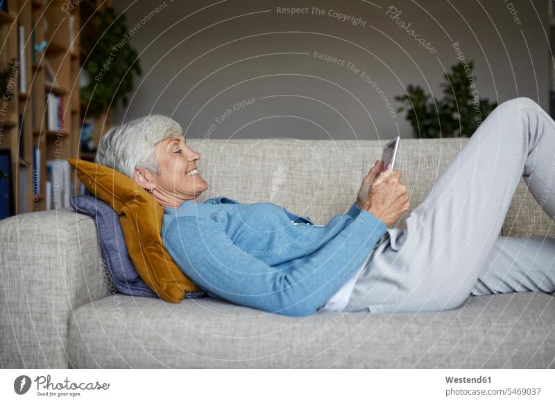 Lächelnde Frau benutzt digitales Tablet, während sie zu Hause auf dem Sofa liegt Farbaufnahme Farbe Farbfoto Farbphoto Innenaufnahme Innenaufnahmen innen